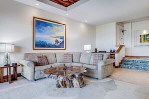 A living room with a sectional sofa, a coffee table made of wood, a painting on the wall, and a staircase leading to another level.