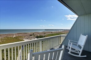 A balcony with a white rocking chair, overlooking a beach with a pier in the distance.