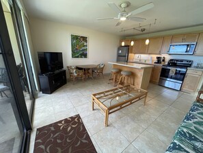 A modern kitchen with a central island, a dining area with wicker furniture, and a TV in the living room.