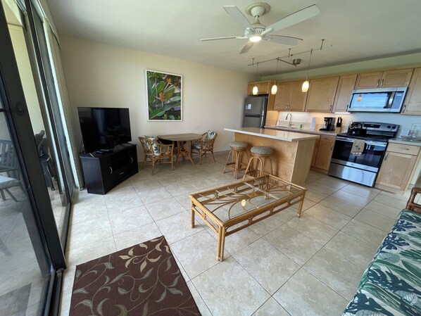 A modern kitchen with a central island, a dining area with wicker furniture, and a TV in the living room. - Waiohuli Beach Hale #D-127 Direct Oceanfront Complex in Kihei, Steps to Beach (Kihei)