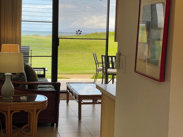 A room with a view of a grassy field and a body of water. There is a table, a chair, and a sofa with cushions. - Waiohuli Beach Hale #D-127 Direct Oceanfront Complex in Kihei, Steps to Beach (Kihei)