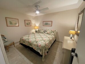 A bedroom with a bed, a chair, a nightstand, a lamp, a ceiling fan, and framed pictures on the wall.