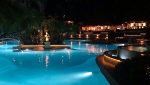 Une piscine éclairée, utilisée la nuit, avec des bordures illuminées et un palmier au premier plan.