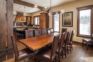 Uma sala de jantar espaçosa com uma grande mesa de madeira e cadeiras de couro, uma cozinha moderna ao fundo e uma janela ampla com vista para árvores.