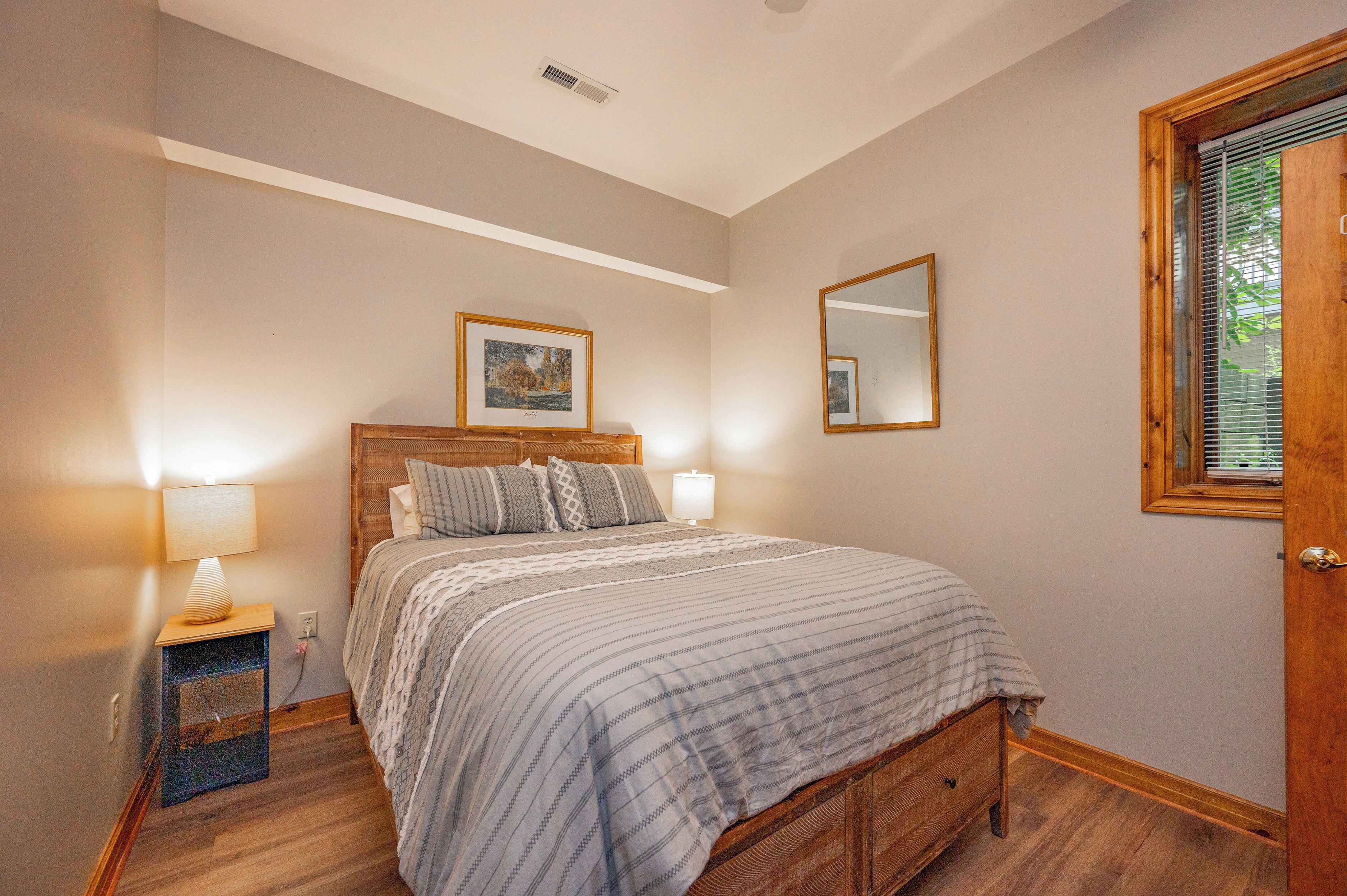 A bedroom with a wooden bed, bedside lamps, a framed picture on the wall, and a window with blinds.