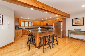 A modern kitchen with wooden cabinets, a central island with stools, stainless steel appliances, and a wooden ceiling beam. - Magical Lake Escape w/ Dock Slip & Game Room (Oakland)