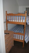 Een stapelbed met houten frame en blauw gestreepte lakens. Er staat een dressoir met lades en een afstandsbediening erop.