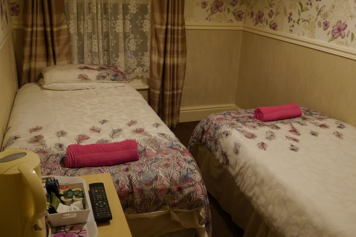 Une chambre d’hôtel avec deux lits simples, chacun recouvert d’une couvre-lit à motifs floraux et agrémenté d’une serviette rose pliée. Une table de chevet en bois porte une lampe jaune, une télécommande et un petit récipient blanc contenant des articles de toilette.