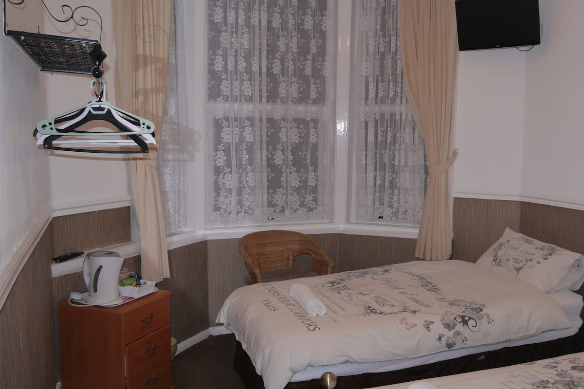 Une chambre à coucher avec un lit, une chaise, une commode, une fenêtre avec des rideaux et un téléviseur fixé au mur.