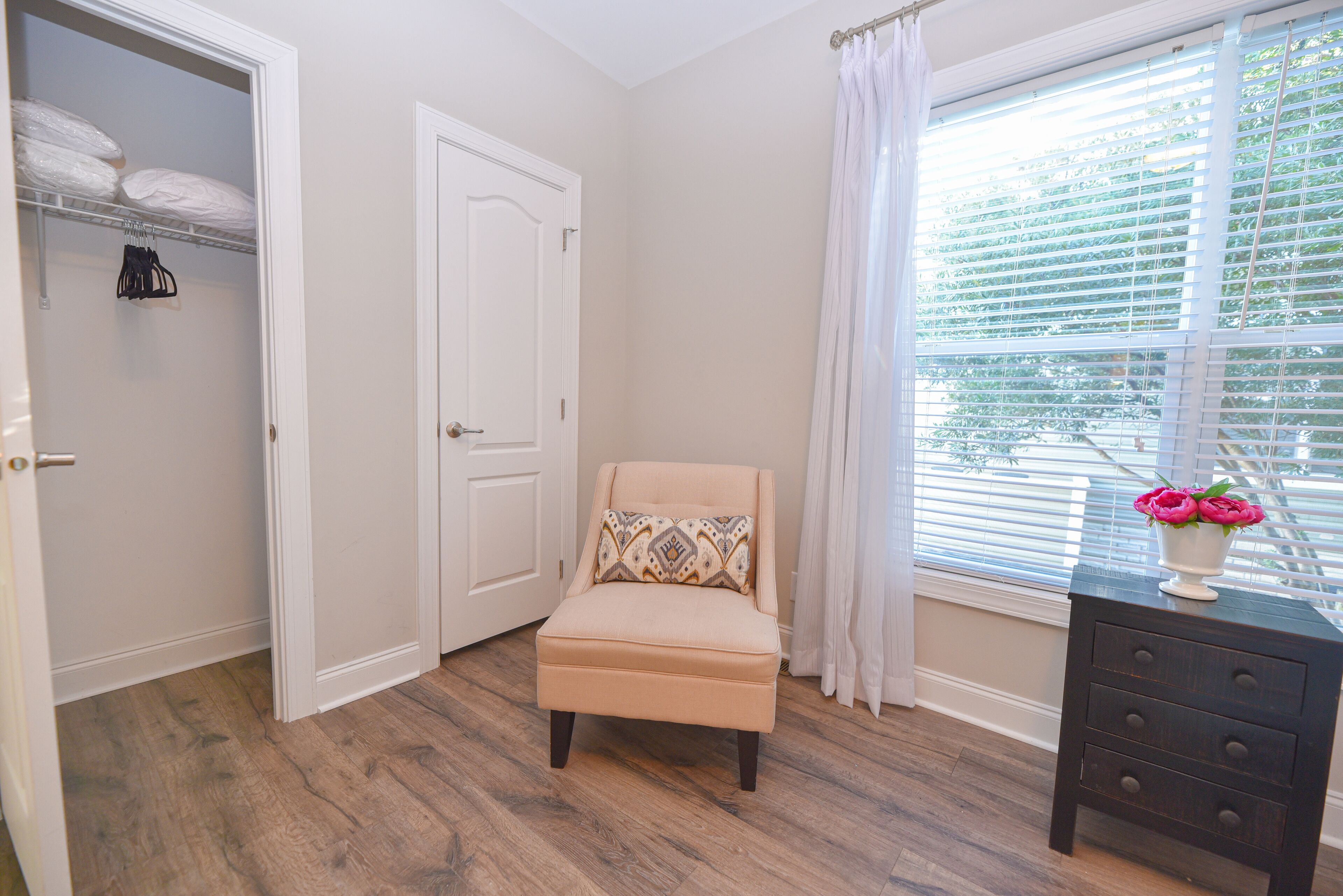 Een kamer met een beige fauteuil, een houten vloer, een witte deur, een raam met zonwering en een dressoir met lades.