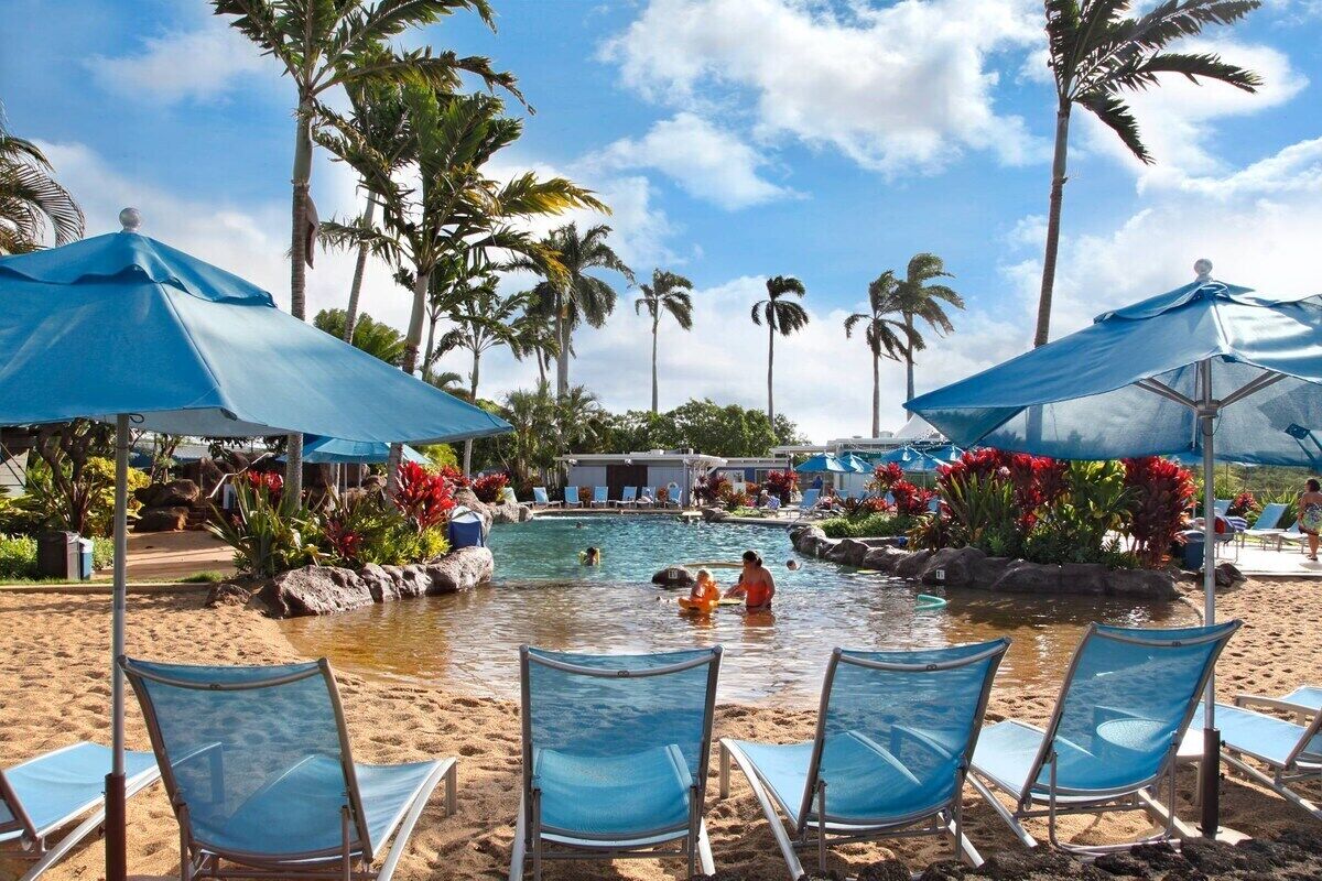 A poolside area with lounge chairs and umbrellas, surrounded by palm trees and tropical plants.