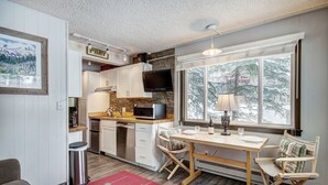 A compact kitchen with a microwave, stove, and sink, a dining area with a table and chairs, and a large window with a view of trees. - Remodeled Corner Unit with Lots of Light! Walk to Town | PM4A (Breckenridge)