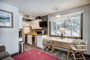 A compact kitchen with a microwave, stove, and sink, a dining area with a table and chairs, and a large window with a view of trees.