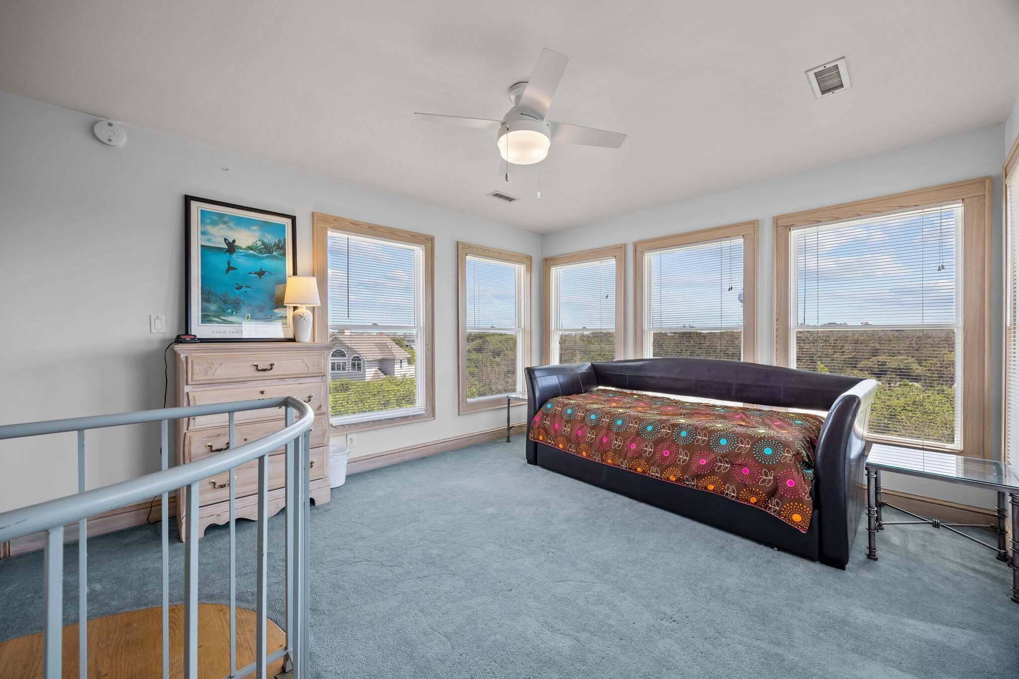 A bedroom with a bed, a ceiling fan, a dresser, a lamp, and large windows.