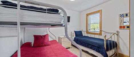 Een slaapzaal met stapelbedden, een klein nachtkastje, een raam met jaloezieën en een aan de muur bevestigde televisie.