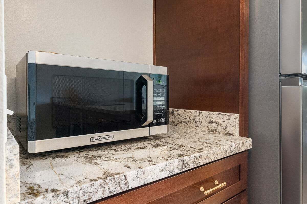 A kitchen with a microwave on a granite countertop, a wooden cabinet, and a stainless steel refrigerator.