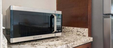 A kitchen with a microwave on a granite countertop, a wooden cabinet, and a stainless steel refrigerator.