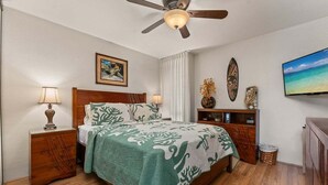 Ein Schlafzimmer mit einem Bett, Holznachttischen, einem Deckenventilator, einem Fernseher und einem an der Wand befestigten Regal mit Dekorationen.
