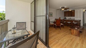 Ein Balkon mit Glastisch und Stühlen, eine Glasschiebetür zu einem Küchenbereich mit Holzboden, einem Deckenventilator und einer Bar.