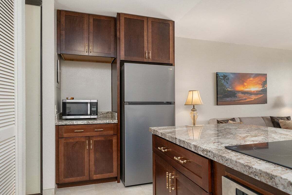 A kitchen with wooden cabinets, a stainless steel refrigerator, a microwave, a granite countertop, and a painting of a sunset.
