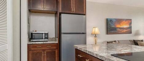 A kitchen with wooden cabinets, a stainless steel refrigerator, a microwave, a granite countertop, and a painting of a sunset.