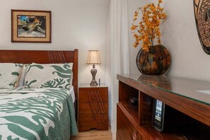 Ein ordentlich bezogenes Bett mit einem gemusterten Überwurf, eine Nachttischlampe, eine dekorative Vase mit Blumen, ein gerahmtes Bild an der Wand und ein Regal mit einer Uhr.