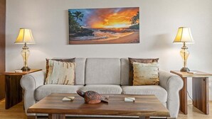 A living room with a sofa, two side tables with lamps, a coffee table with a decorative item, and a painting of a sunset over the ocean.