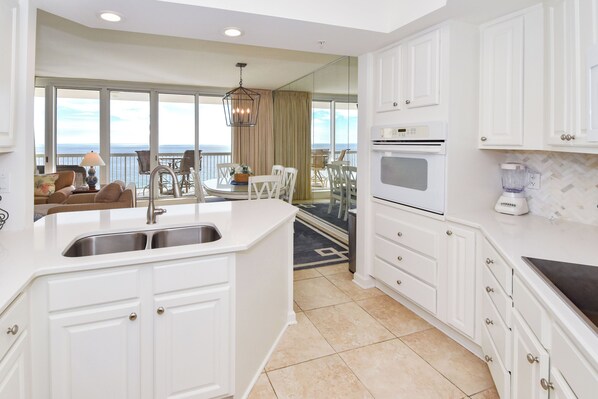 A modern kitchen with white cabinetry, a built-in oven, and a large island with a sink. There is a dining area with a table and chairs, and a view of the ocean through large windows. - Warm, welcoming beachfront condo w/private balcony! ☀️ (Destin)