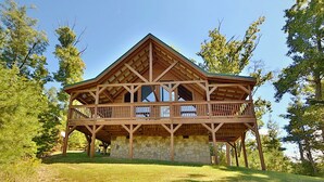 A wooden cabin with a stone foundation, a balcony, and a clear view of the surrounding trees. - Secluded | Pet Friendly | Pool Table | Arcade | Hot Tub | WiFi (SEVIERVILLE)