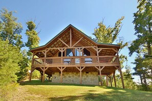 A wooden cabin with a stone foundation, a balcony, and a clear view of the surrounding trees. - Secluded | Pet Friendly | Pool Table | Arcade | Hot Tub | WiFi (SEVIERVILLE)
