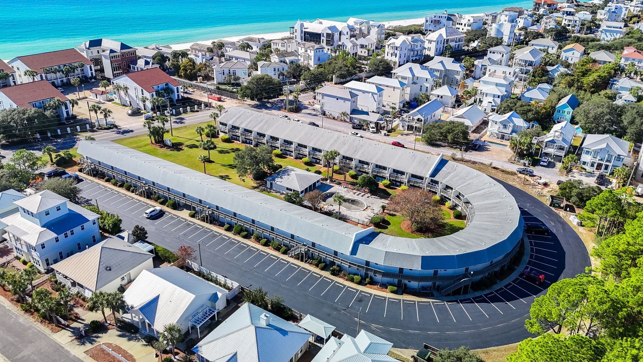 Vista aérea de una zona residencial costera con un gran edificio curvo, probablemente un hotel o resort, que cuenta con un estacionamiento y una playa al fondo.