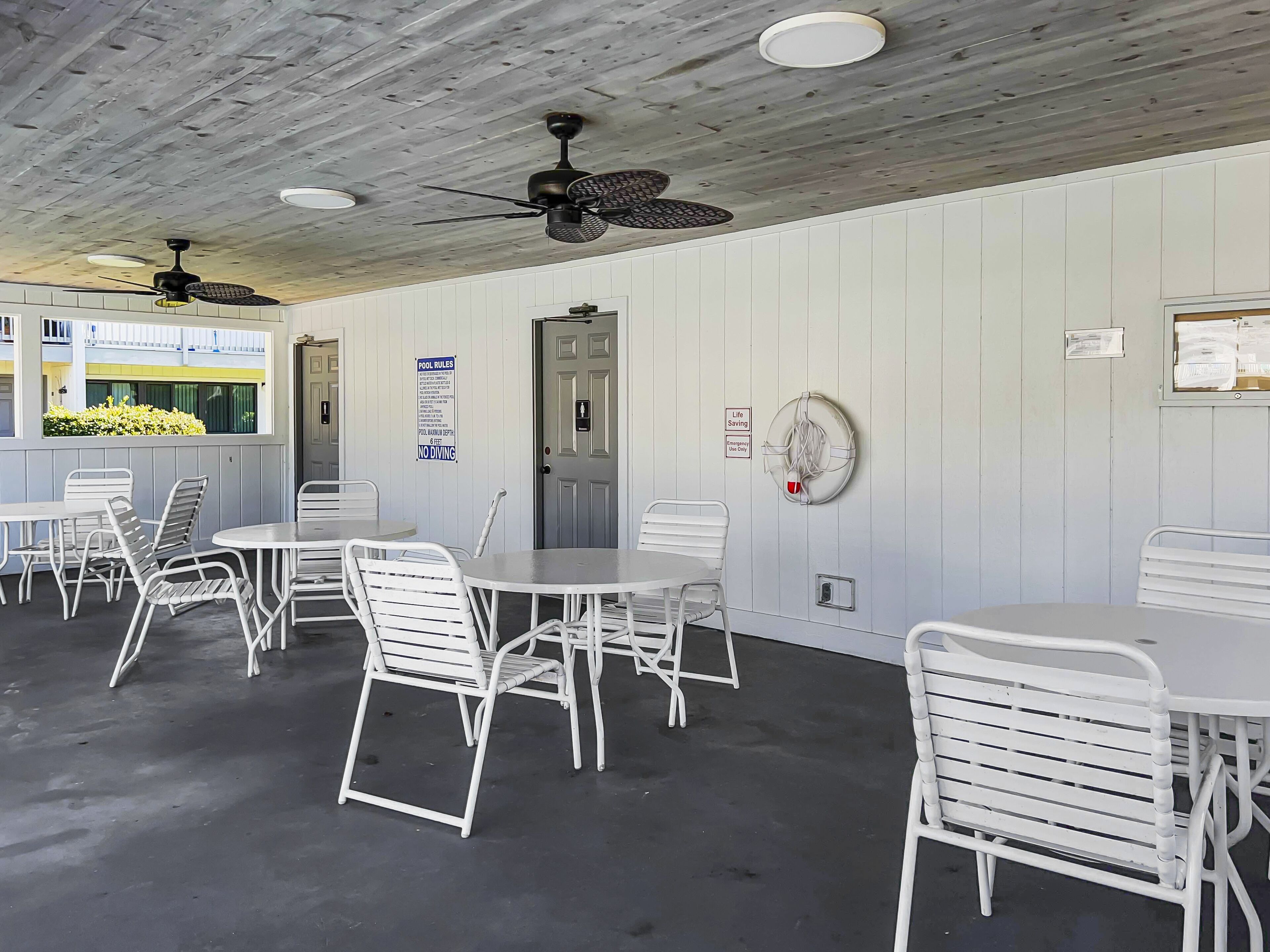 A room with white metal chairs and tables, a ceiling fan, and a door with a sign indicating pool rules.