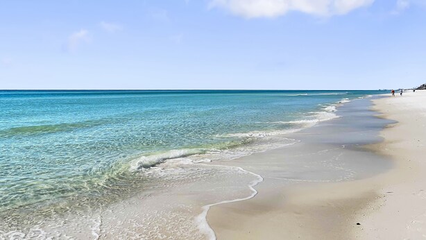 Ein Strand mit klarem türkisblauem Wasser, einem sandigen Ufer und einigen Spaziergängern.