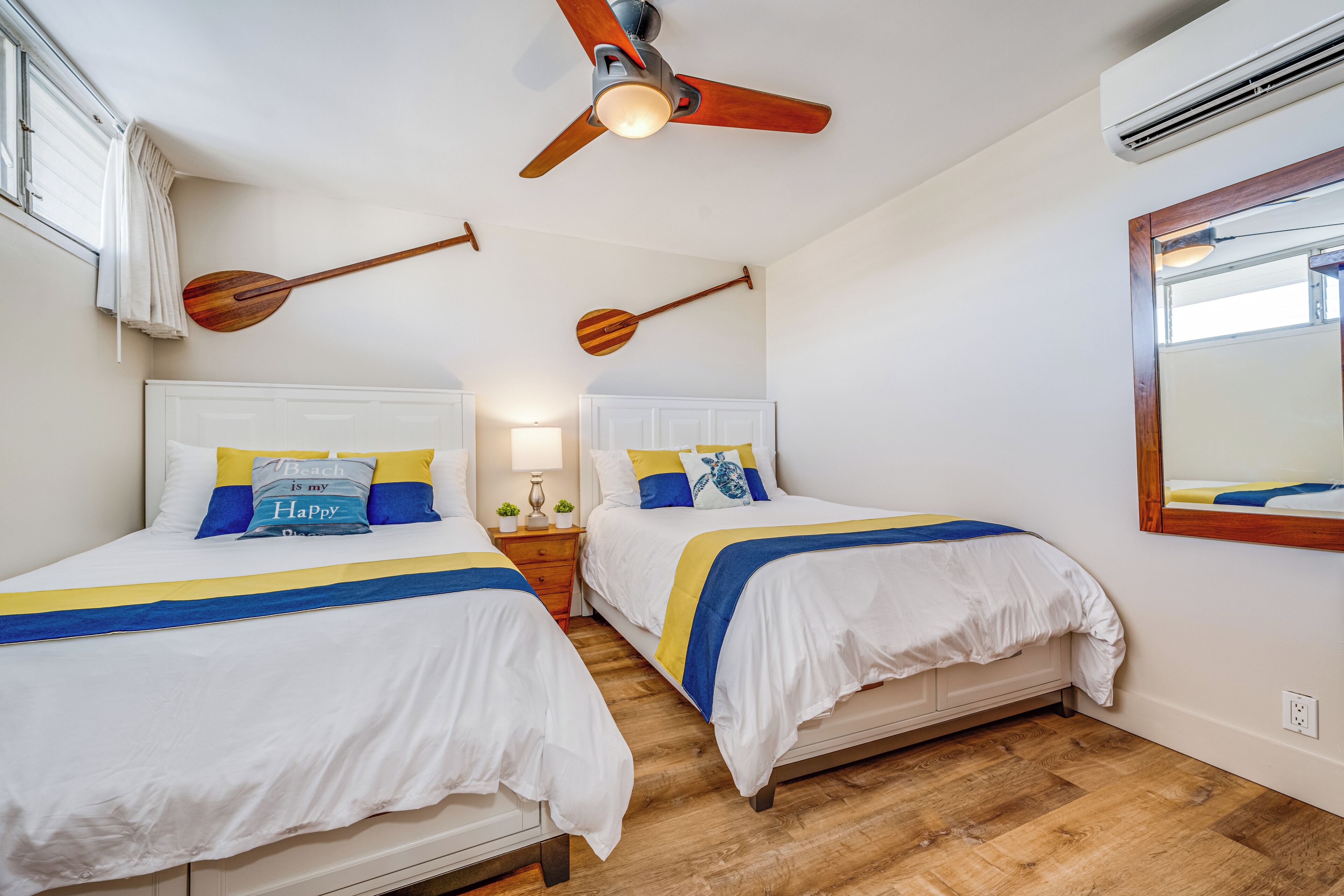A hotel room with two beds, wooden floors, a ceiling fan, and a large mirror.