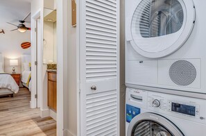 A laundry room with a stacked washer and dryer, a ceiling fan, and a door leading to a bedroom.