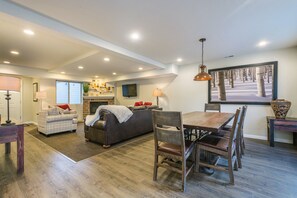 Un salon spacieux avec une cheminée, un coin repas avec des meubles en bois et une grande fenêtre donnant sur les arbres.
