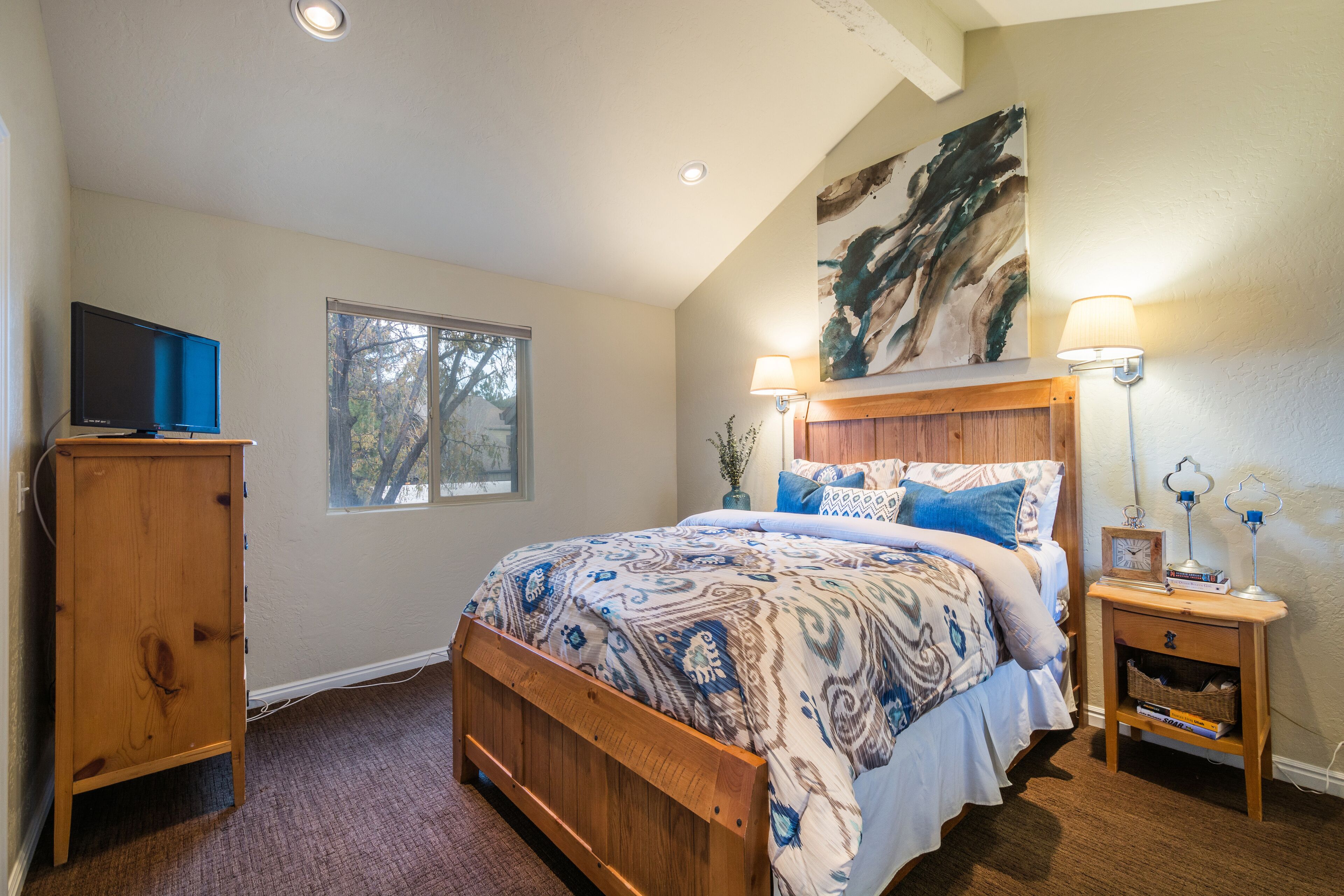 A bedroom with a wooden bed, a TV on a stand, a window, and a painting on the wall.