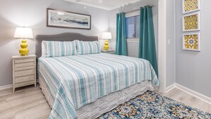 Un dormitorio con una cama, dos mesitas de noche con lámparas, un ventilador de techo, una ventana con cortinas y cuadros en la pared.