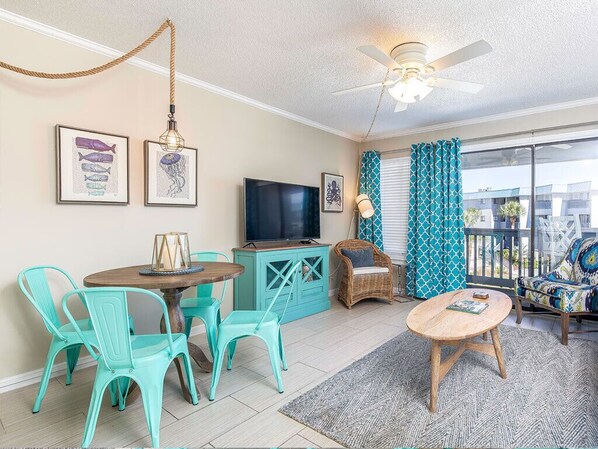 A living room with teal chairs, a wooden coffee table, a TV, and a ceiling fan. - Beach Racquet B219 | Steps to the Beach + Pool! (Tybee Island)