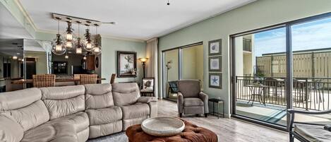 A spacious living room with a large sectional sofa, a brown ottoman, and a dining area with a glass balustrade.