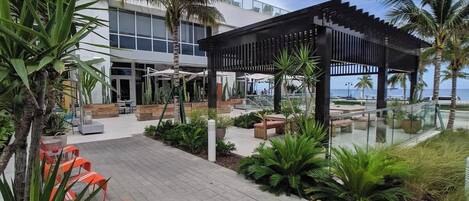 A modern building with a glass facade, outdoor seating, and a palm tree-lined patio.