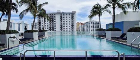 Una piscina con barrera de cristal, palmeras y edificios altos al fondo.