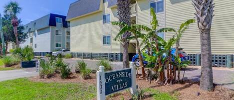 Señal de Oceanside Villas, un edificio de varios pisos con fachada amarillo claro, palmeras y cielo despejado.