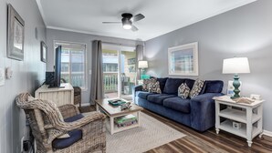 A living room with a blue sofa, wicker chair, and a ceiling fan. - Modern Condo, Pool, Close to Beach, Restaurants & Shops - 101A Oceanside (Isle Of Palms)