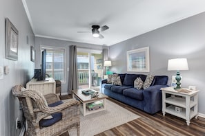 A living room with a blue sofa, wicker chair, and a ceiling fan. - Modern Condo, Pool, Close to Beach, Restaurants & Shops - 101A Oceanside (Isle Of Palms)