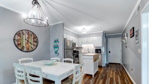 A modern kitchen with white cabinetry, a central island, and a dining area with white chairs and a table. - Modern Condo, Pool, Close to Beach, Restaurants & Shops - 101A Oceanside (Isle Of Palms)