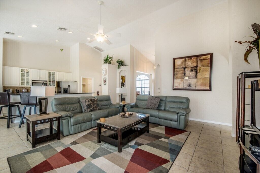 A spacious living room with a sectional sofa, a coffee table, and a patterned rug. A kitchen is visible in the background with white cabinets and a dining area.