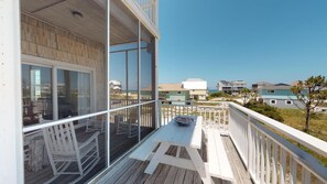 Une terrasse en bois avec une balustrade blanche, une table entourée de chaises et, en arrière-plan, une vue sur la plage et d’autres bâtiments.