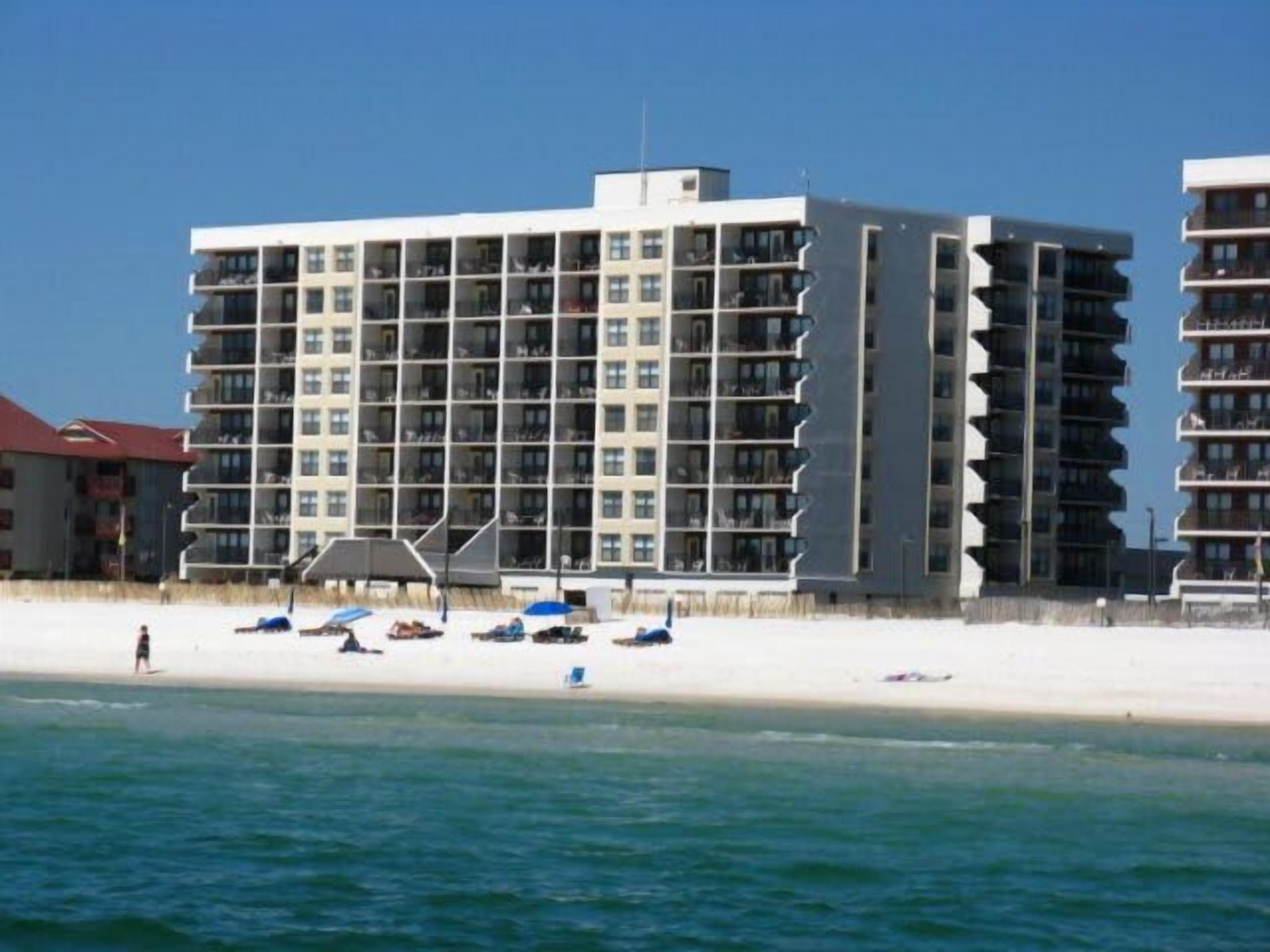 Ein mehrstöckiges Hotel mit Balkonen in Strandnähe, mit Sonnenschirmen und Liegestühlen.