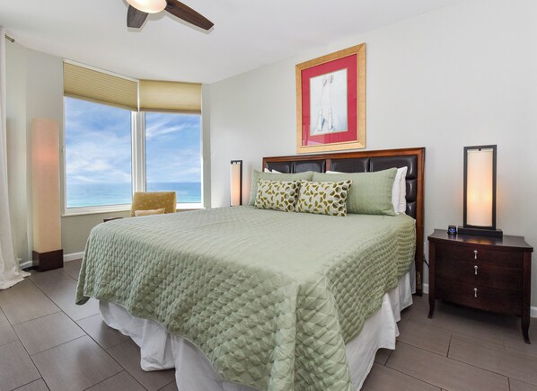 A hotel room with a large bed, a view of the ocean, a ceiling fan, and a framed artwork on the wall.